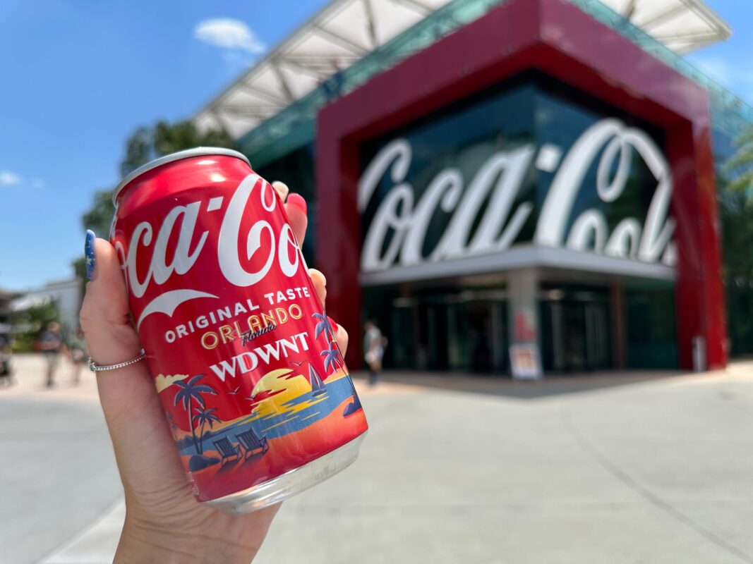 A hand holds a Coca-Cola can with an Orlando WDWNT design in front of a building with large Coca-Cola signage.