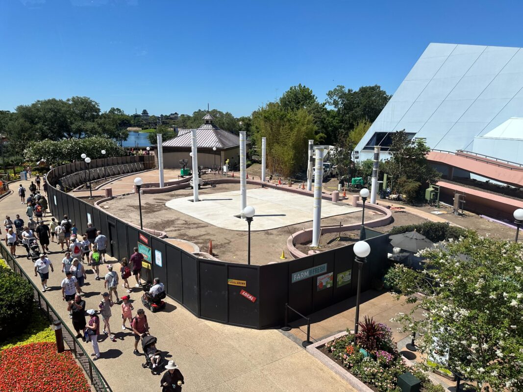 Many people are walking on a pathway beside flex space construction site with fenced-off areas and building materials. Several vertical columns are in progress, and various landscaping is in the vicinity.
