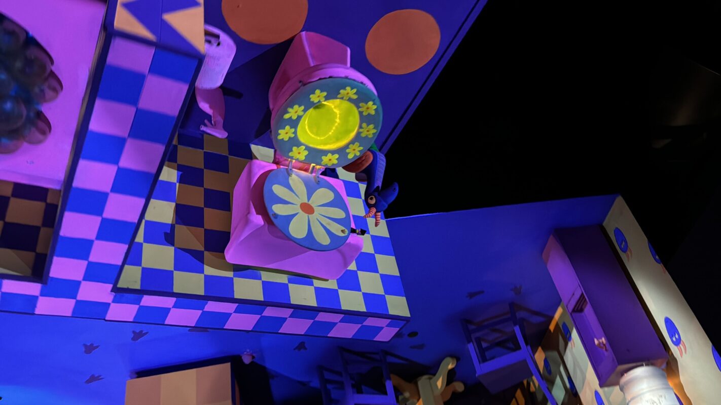 A colorful, whimsical scene from Journey Into Imagination with Figment featuring vibrant, geometric shapes and patterns, including a large, flower-decorated upside-down toilet on a checkered floor. The setting is brightly lit and playful.