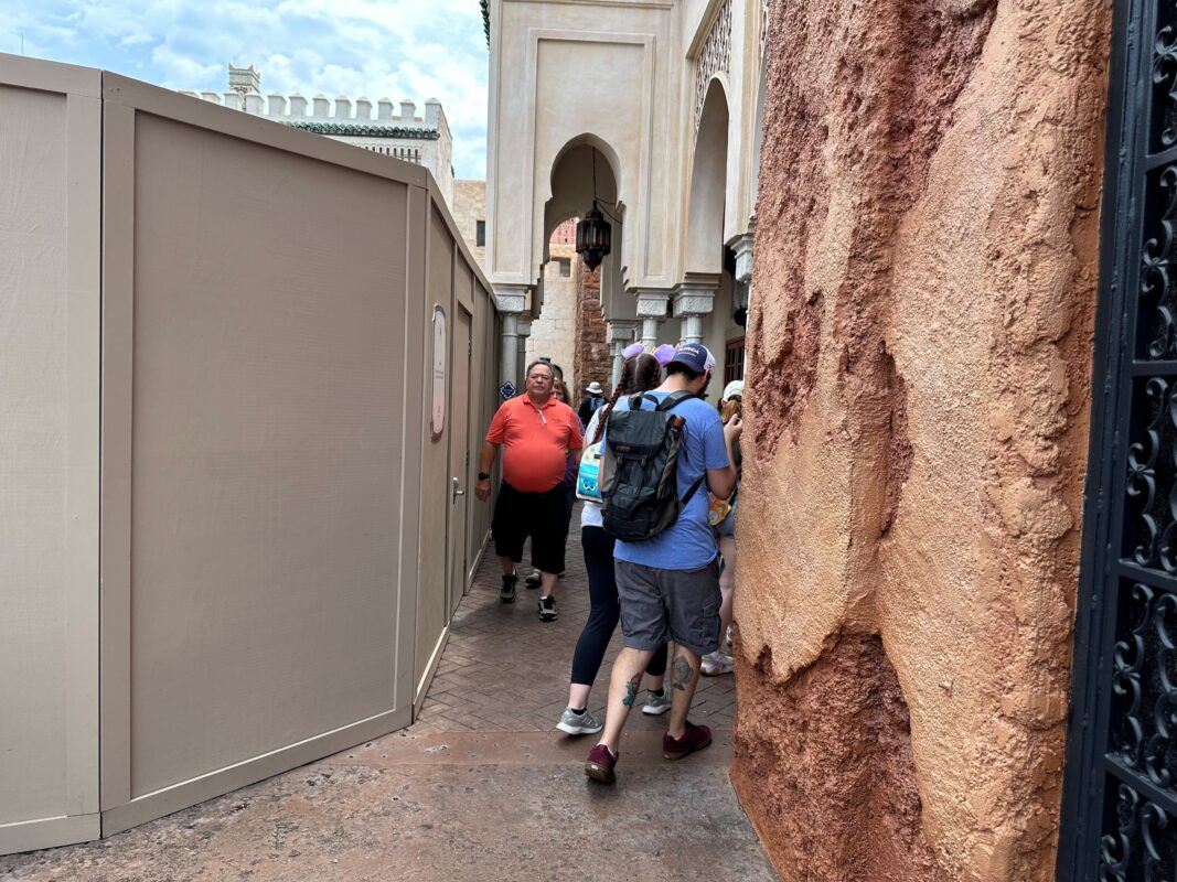 People walking through a narrow passageway in an outdoor setting with construction walls on the left and stone walls on the right. A man in an orange shirt leads the group.