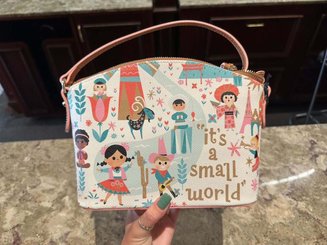 A person holding a small purse with a colorful "it's a small world" theme featuring stylized cartoon children and cultural motifs.