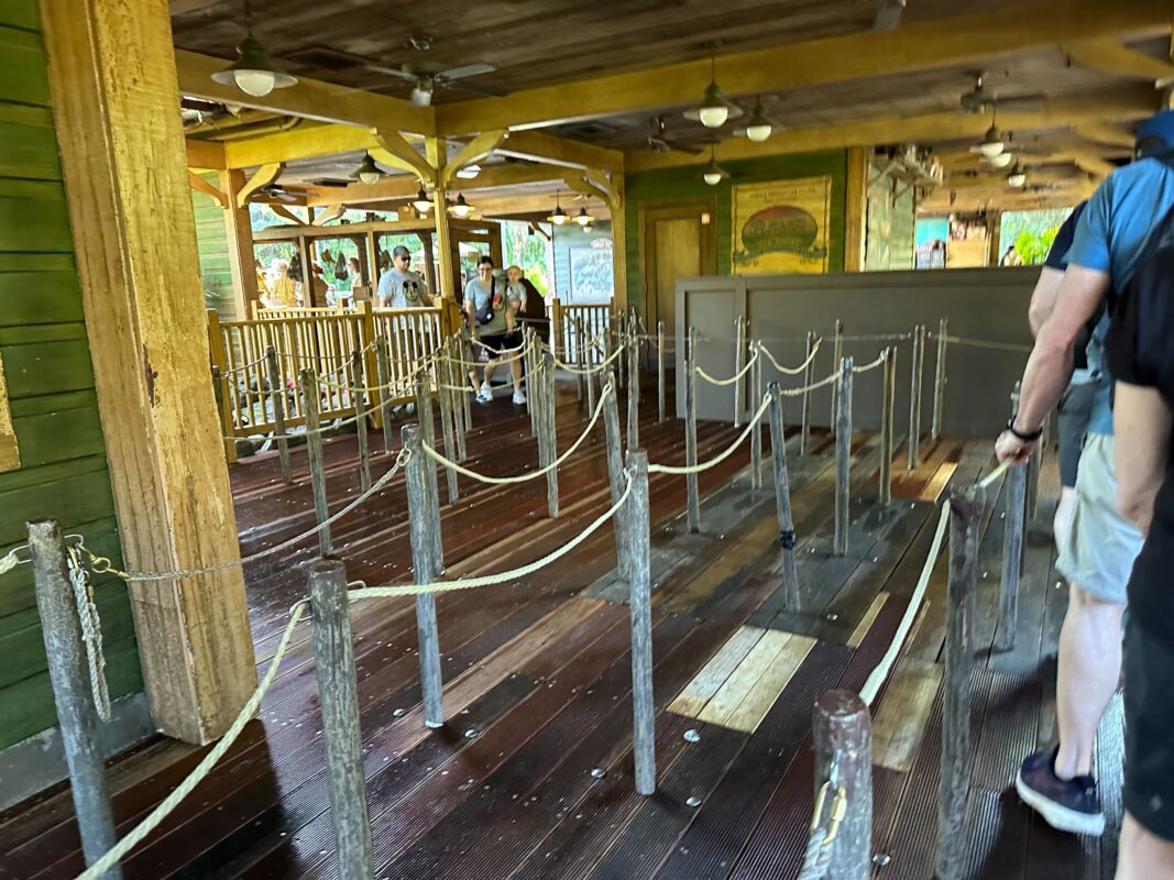 People walking through a rope-lined queue at an amusement park ride station with wooden floors and beams.