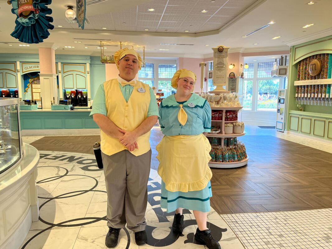 Two employees wearing pastel-colored uniforms and hats stand inside a brightly lit ice cream shop, with various treats displayed in the background.