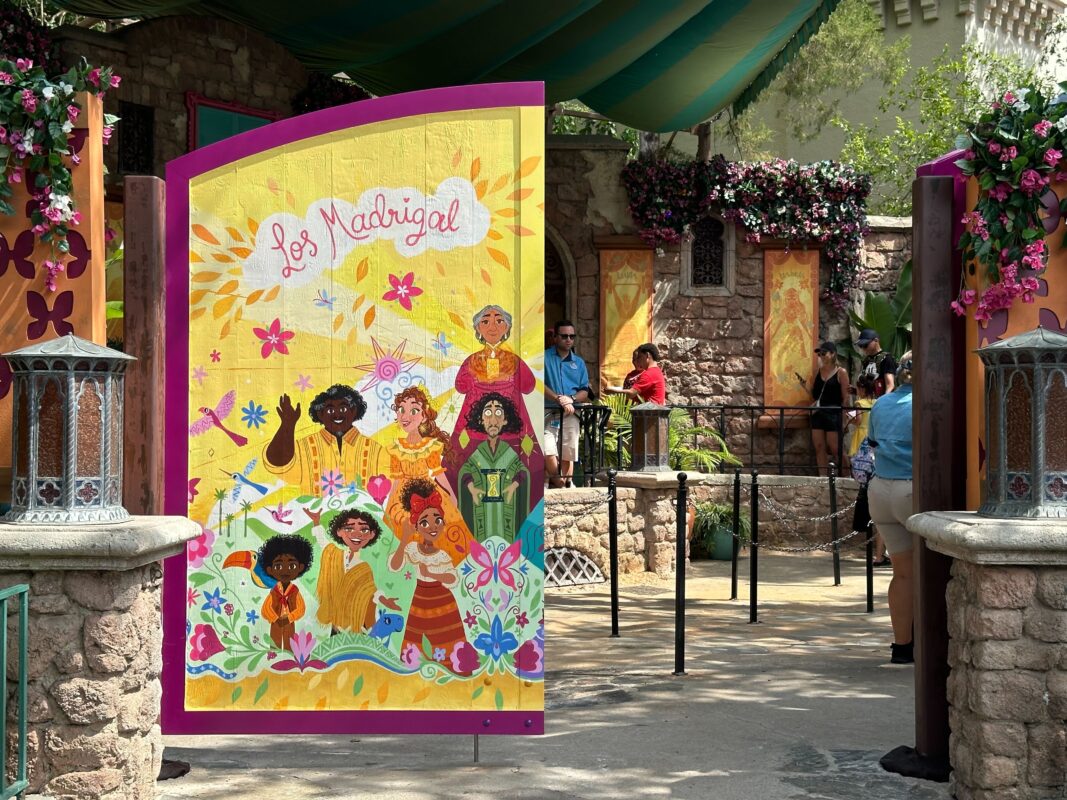 Colorful entrance signage reading "Los Madrigal" featuring illustrated characters, set within a stone and brick surroundings adorned with flowers and lights. Several visitors are visible in the background.