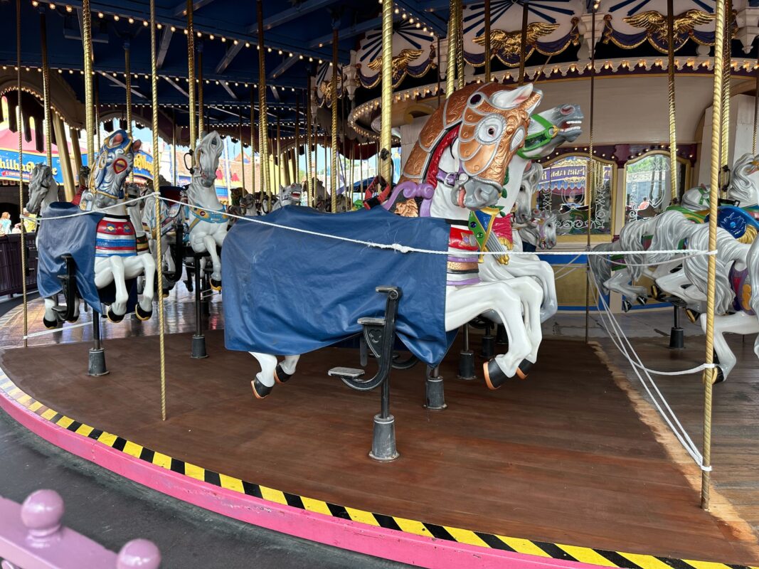 A merry-go-round with ornate, colorful horse figures. Some horses are covered with blue protective sheets. The ride's platform and safety barriers are visible.