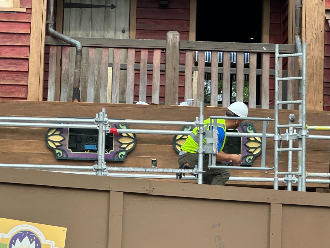 A worker in a high visibility vest and helmet installs wait time signs on a decorative panel at a construction site with scaffolding.