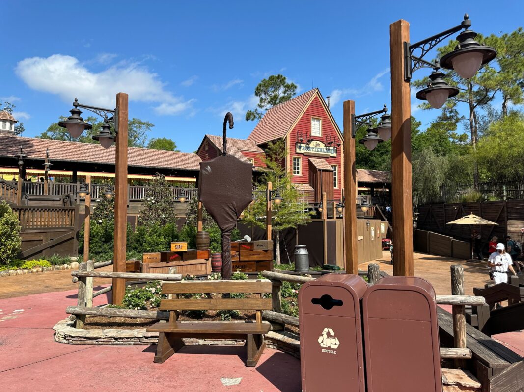 Theme park setting with a rustic bench, lanterns, and a wooden barrel near a trash can with a recycling symbol, overlooked by a red-roofed building and elevated wooden walkway in Tiana's Bayou Adventure.