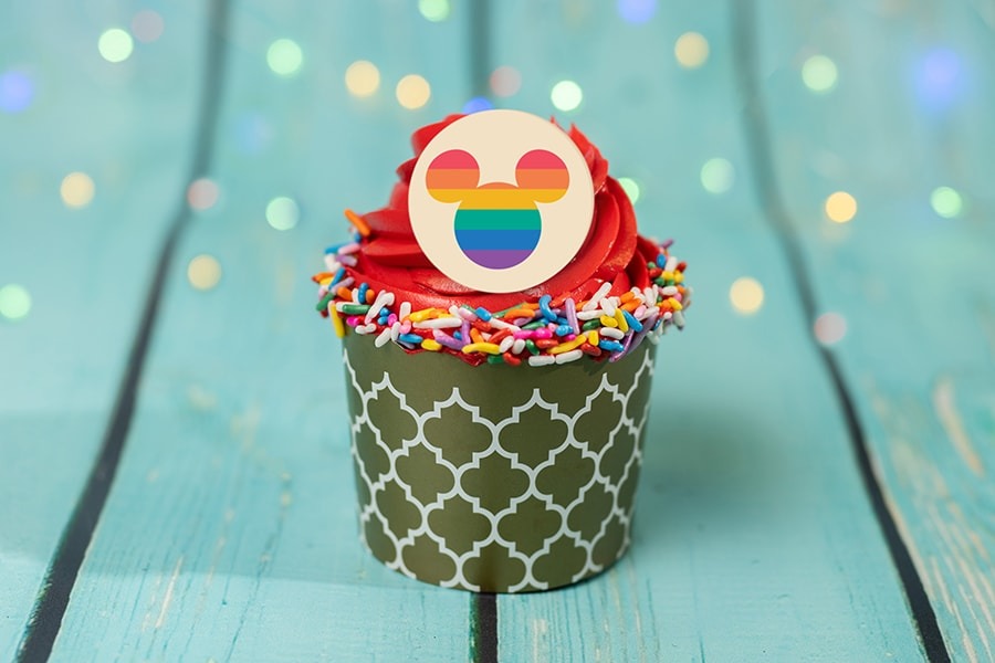 Cupcake with red frosting, rainbow sprinkles, and a circular topper featuring the silhouette of mouse ears in front of a rainbow, on a turquoise wooden surface with a light bokeh background.