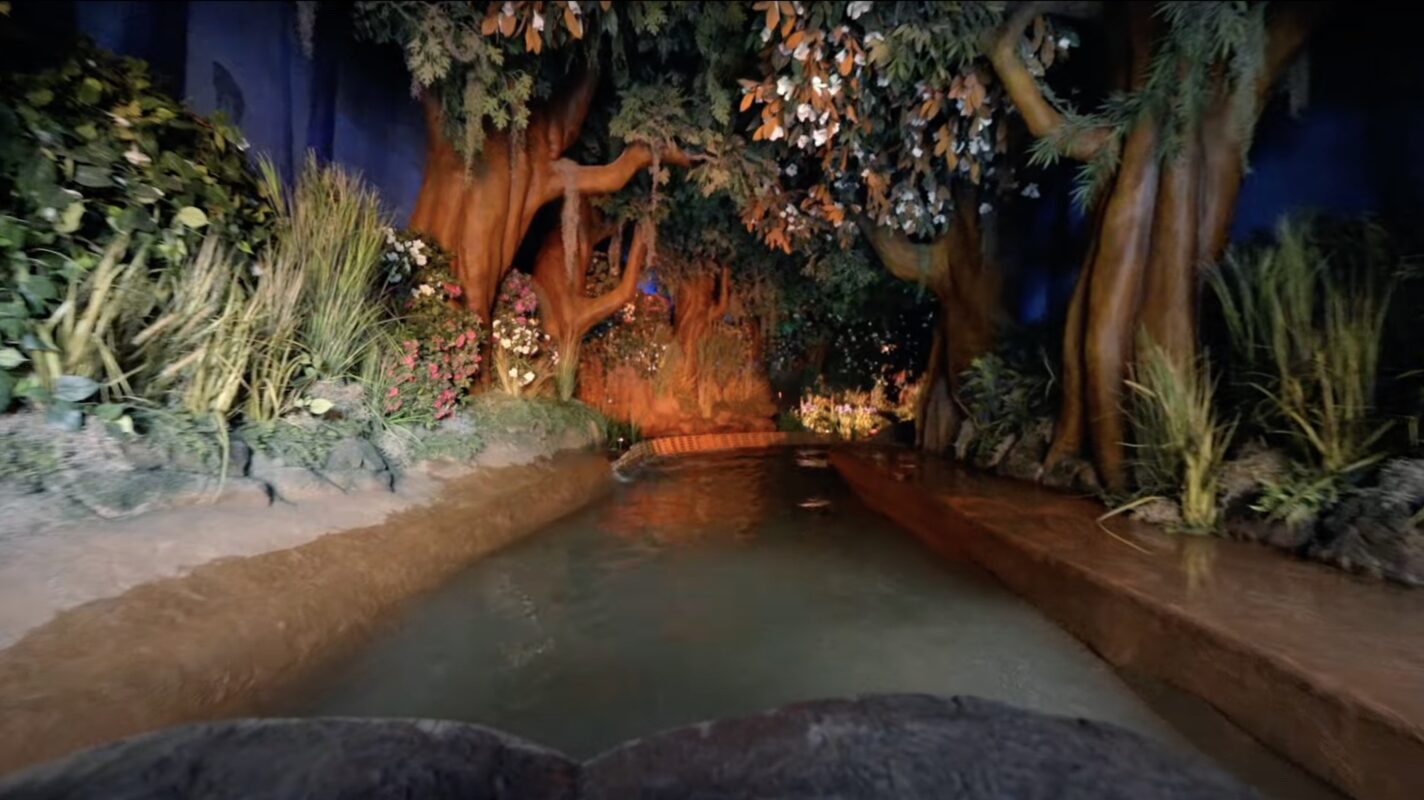 A water ride path surrounded by artificial trees and foliage, evoking a forest scene. The waterway is bordered by low walls and the background features a dimly lit setup.