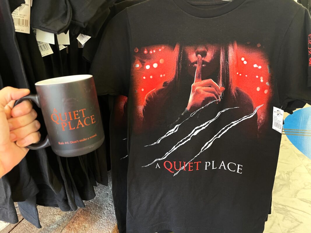 A hand holding a black mug with "A QUIET PLACE" and "Rule #1: Don’t make a sound" text, beside a black t-shirt featuring a hooded figure with claws design labeled "Quiet Place".