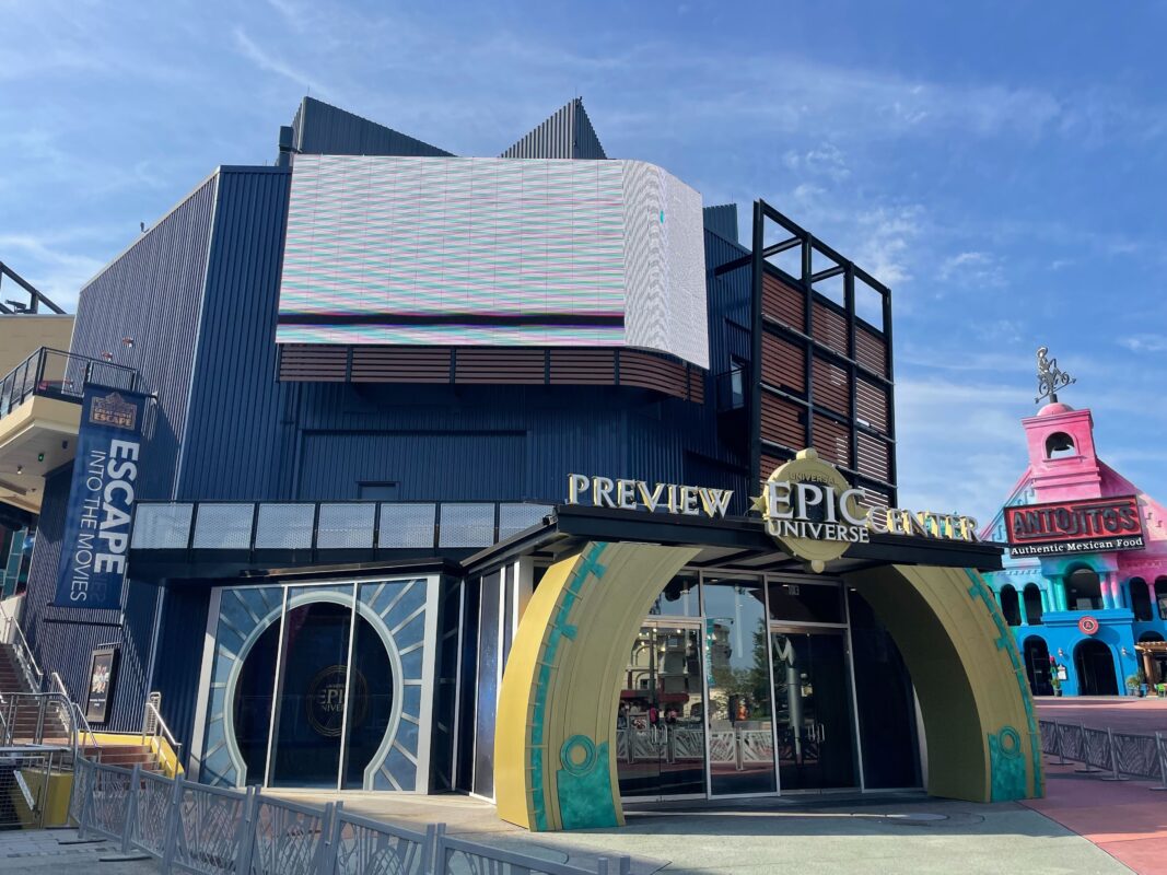 A modern building with a large entrance arch labeled "Preview Epic Universe Center" and a malfunctioning screen above. A sign "Escape into the Movies" is visible, with another colorful building to the right.