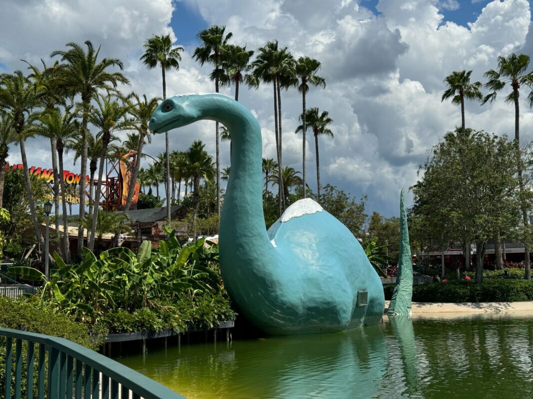 Large blue dinosaur sculpture in water surrounded by lush plants and palm trees under a cloudy sky at a theme park.