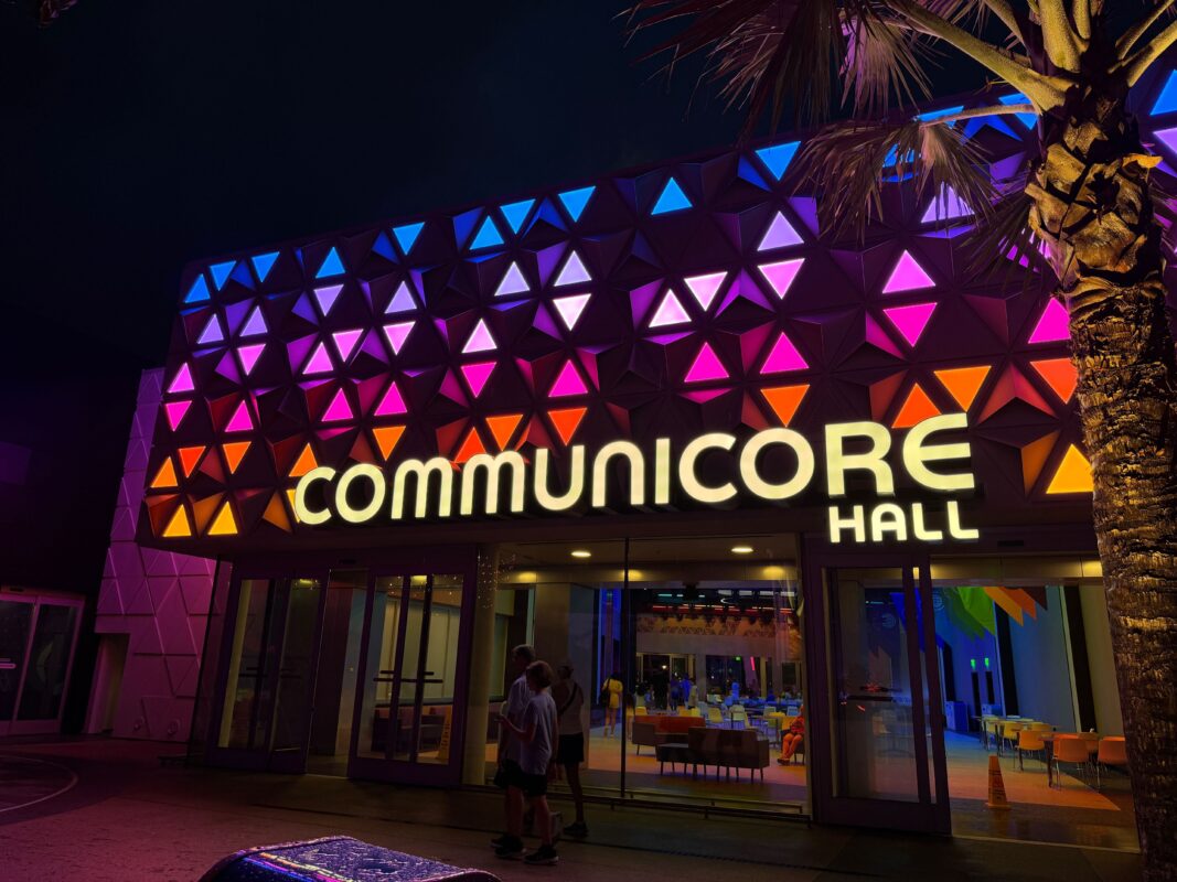 The image shows the exterior of CommuniCore Hall at night, beautifully illuminated with colorful triangular lights. A few people are seen near the entrance, adding a sense of bustling activity.
