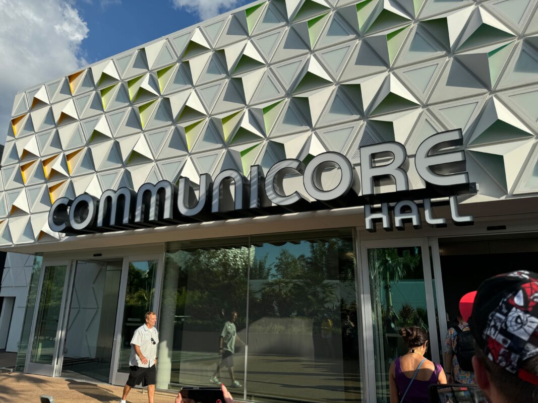 The exterior of CommuniCore Hall features geometric wall designs and large signage, all set under a partly cloudy sky. People are seen walking and standing near the entrance, adding life to the scene around CommuniCore Hall.