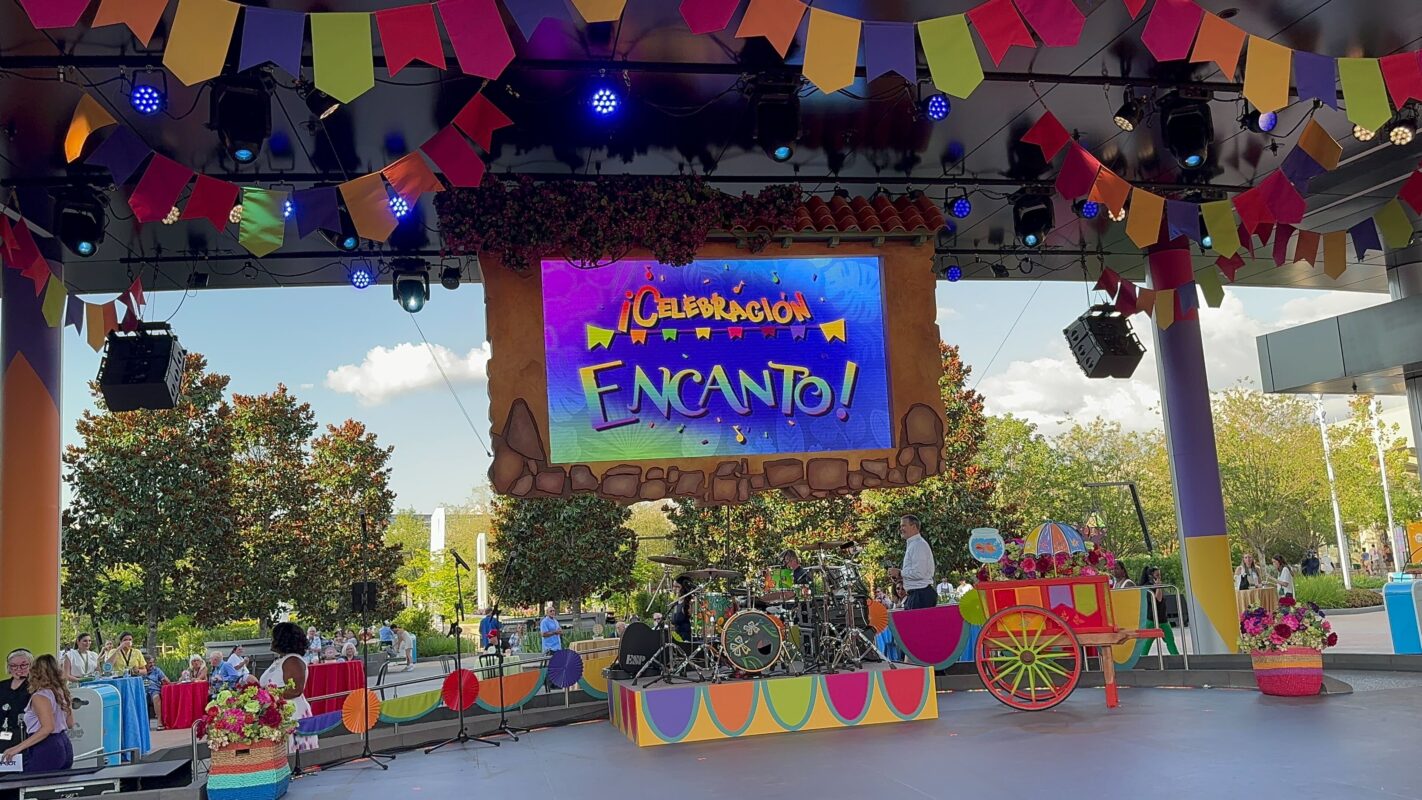 The outdoor stage, adorned with colorful banners and props, features a prominent sign reading "¡Celebración Encanto!" Musical instruments are set up, ready for a lively performance. Trees create a natural backdrop while people excitedly gather around the celebration.