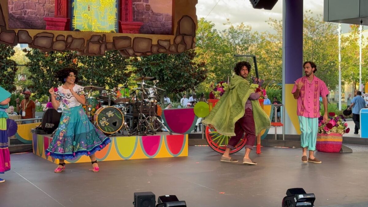 Performers on a colorful stage with a drum set and tropical-themed decorations, including flowers and a bicycle cart, in the background. Some are in costumes; one wrapped in a green cloak. Trees visible behind. The scene radiates the vibrancy of a true celebración encanto.