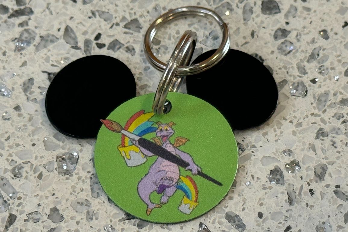Keychain with a circular Figment ID Tag featuring a purple dragon holding a paintbrush with a rainbow, attached to two black Mickey Mouse ear-shaped keys.