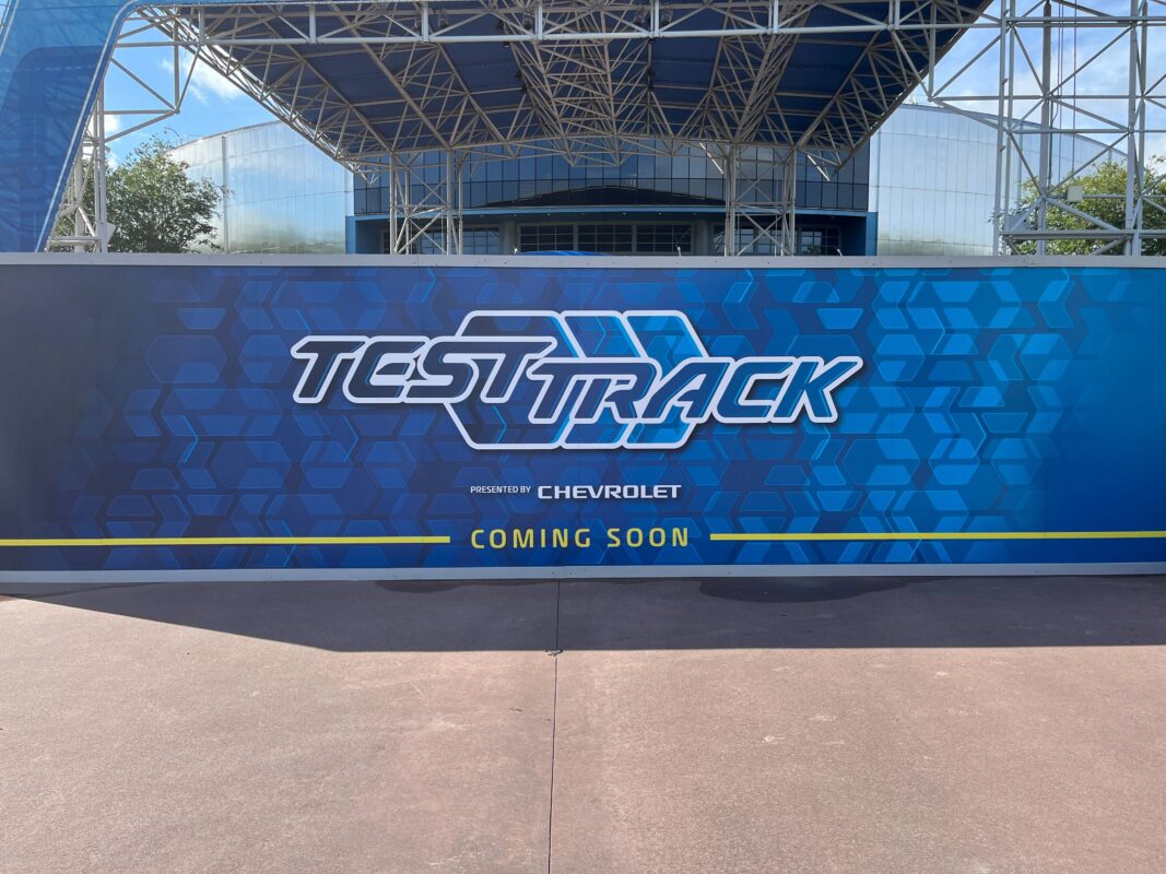 A blue sign with hexagonal patterns reads "Test Track presented by Chevrolet; Coming Soon." It is set against a construction site with a partially visible building and steel structure behind it.