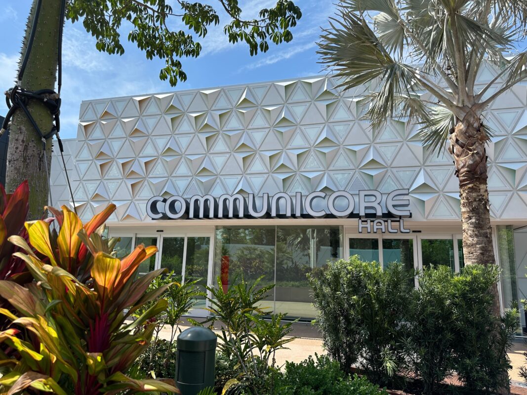 An exterior view of CommuniCore Hall at EPCOT, featuring a geometric-patterned facade with plants in the foreground on a sunny day as the walls come down to reveal its stunning design.