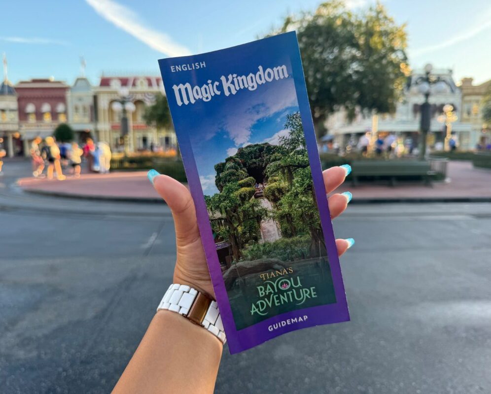 A hand holding a Magic Kingdom guide map featuring "Tiana's Bayou Adventure" on the cover, with a theme park area visible in the background.