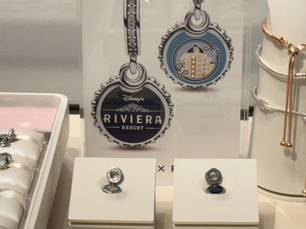 A display featuring jewelry items from Disney's Riviera Resort, including two keychains with round charms, two pairs of earrings on stands, and a Pandora charm bracelet, all showcasing the resort's logo and design motifs.