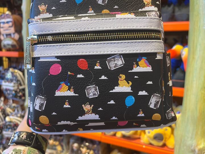 A black and white backpack with a colorful cartoon pattern featuring characters, balloons, and clouds is being displayed in a store. The background shows shelves with various other products.
