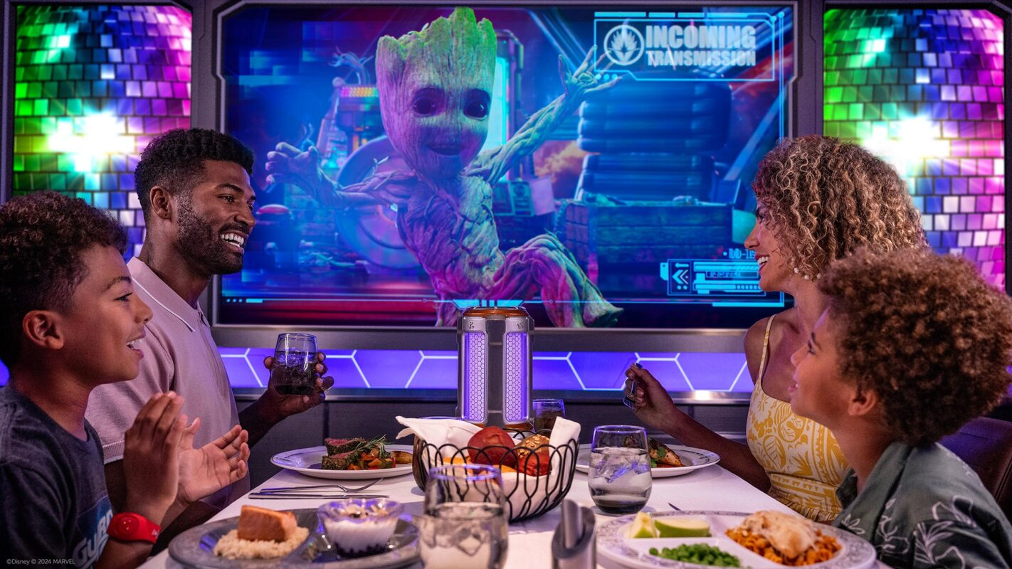 A family of four dines together in the vibrant, brightly lit room of Disney Treasure, immersed in the magic of Worlds of Marvel. As they enjoy their meal, Groot from Guardians of the Galaxy appears on a screen behind them with a cheerful message that reads "Incoming Transmission.