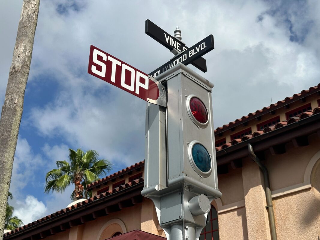 A vintage stoplight with red and green lights stands at the intersection of Vine St. and Hollywood Blvd., against a backdrop of building and palm trees under a partly cloudy sky.
