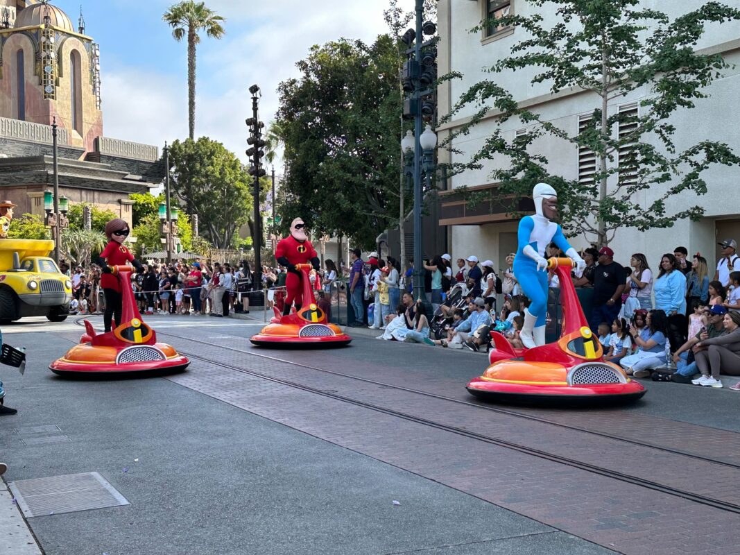 During the Pixar Pals Celebration, three people in "The Incredibles" costumes ride red hovercraft-like vehicles down a street lined with excited spectators. Buildings and trees create a vibrant backdrop at Disney California Adventure.