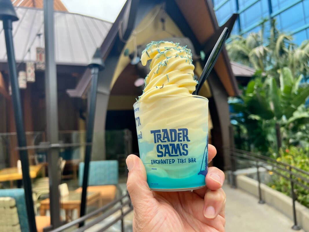 A hand holds a soft serve ice cream in a Trader Sam's Enchanted Tiki Bar cup, with a building and outdoor seating area in the background.