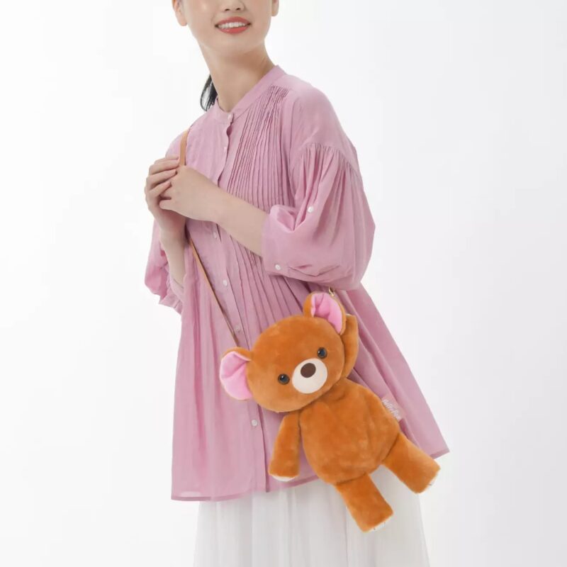A person in a pink shirt and white skirt is holding a brown teddy bear shoulder bag against a plain white background.