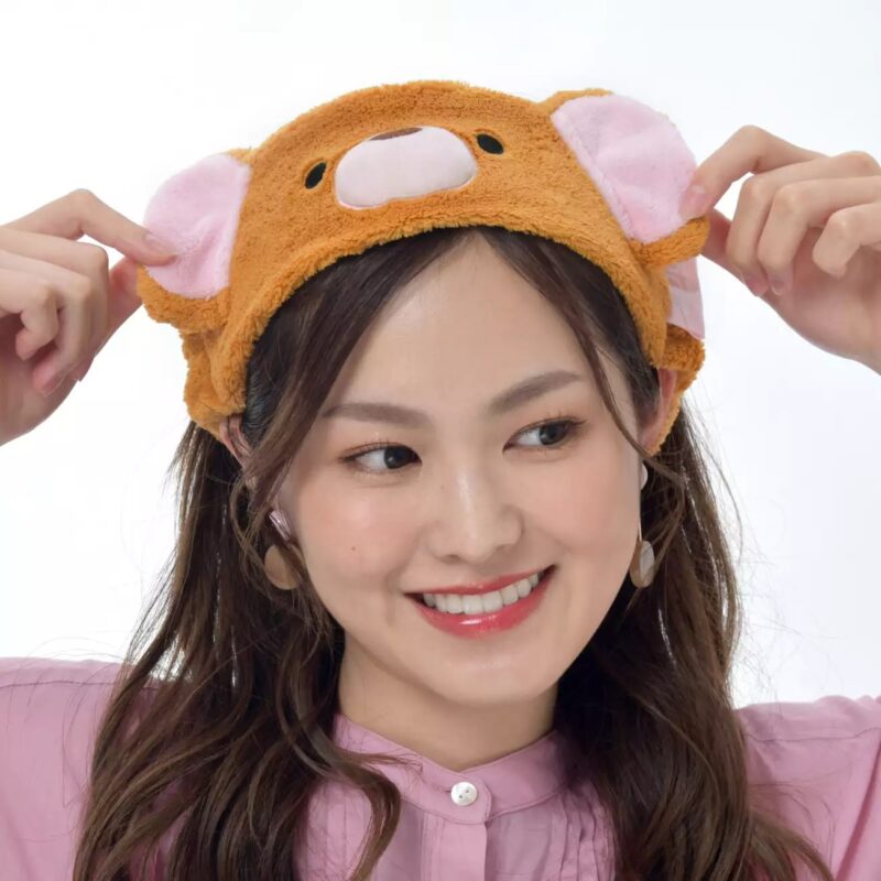 A woman wearing a brown headband with bear ears smiles and holds the ears with her hands. She has long brown hair and is dressed in a pink top.