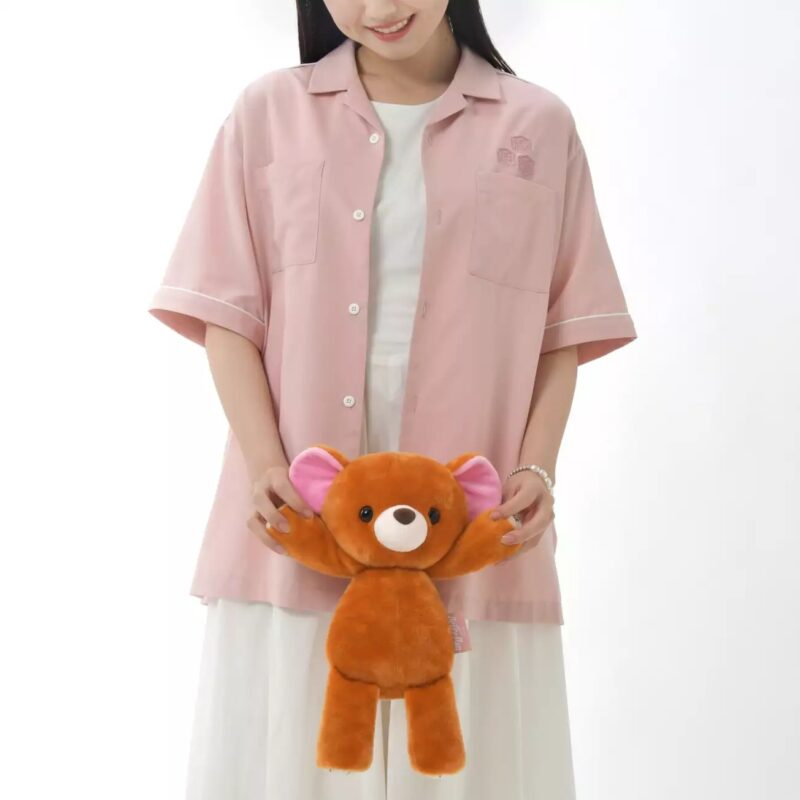 A person wearing a pink short-sleeve shirt and white skirt holds a small brown teddy bear with outstretched arms.