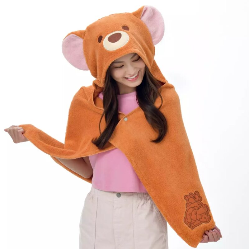 A woman wearing a brown hooded blanket with bear ears and face, smiling and looking down. She is dressed in a pink shirt and beige skirt.