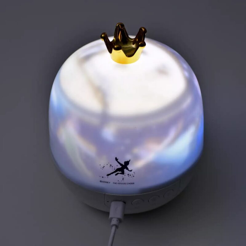A small nightlight with a crown-shaped top emits a soft glow. The light features Disney branding and a silhouette of Tinker Bell printed on its side. It is connected via a USB cable.