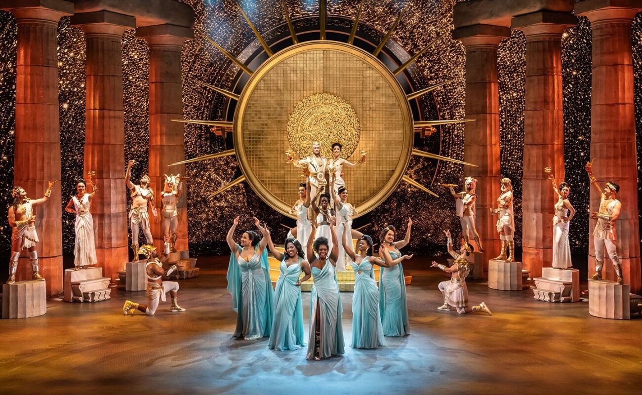 A Broadway musical will bring to life a theatrical performance on a big stage, with actors in ancient-themed costumes, featuring a large golden circular backdrop, columns, and dancers in the foreground wearing light blue dresses.