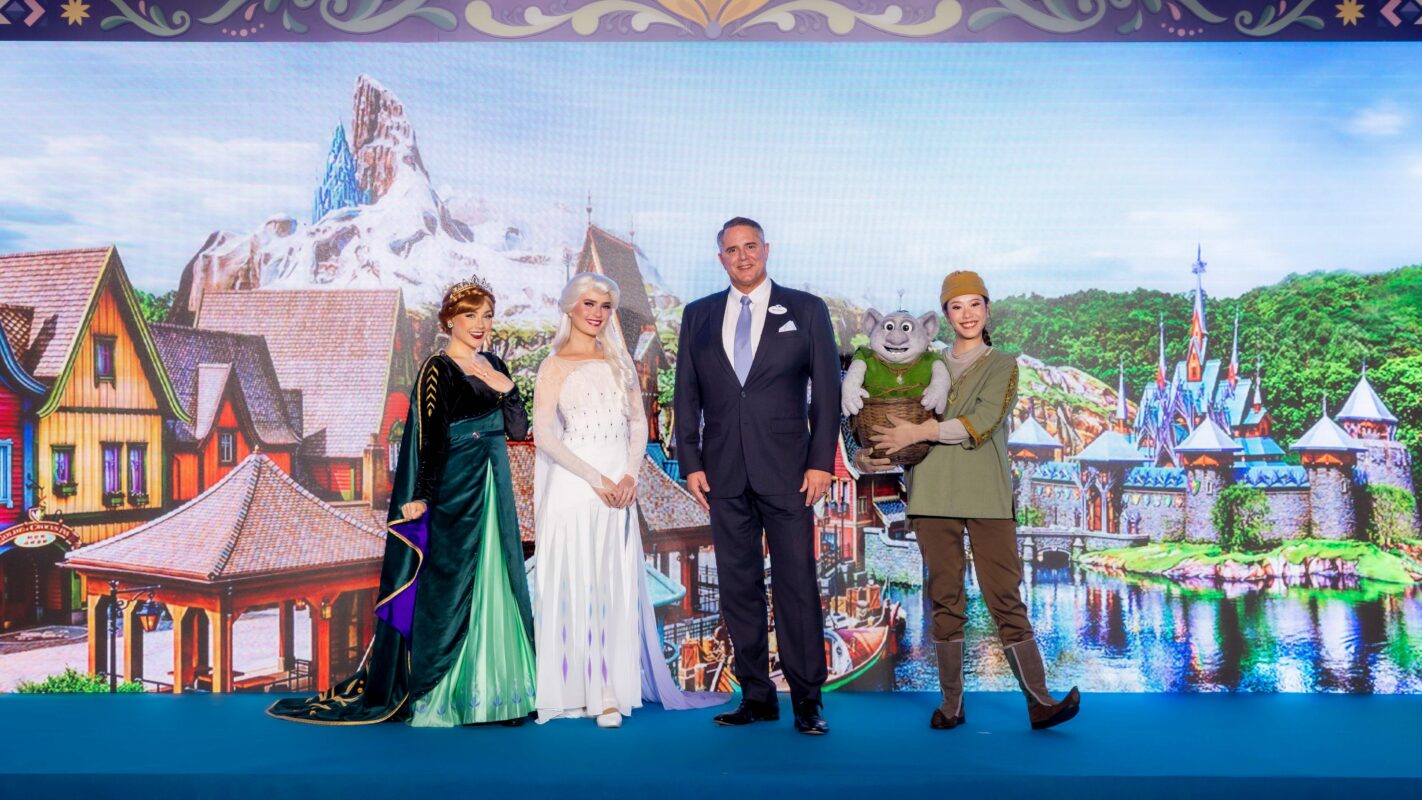 A group of people, including individuals in fantasy costumes and one in formal attire, stand in front of a backdrop featuring an animated village and mountain landscape.