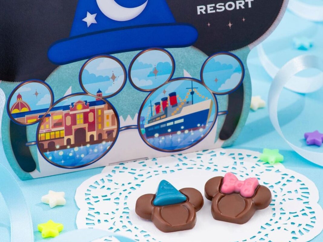 A colorful themed box with a wizard hat, glasses, and ears illustration is displayed. Two Mickey-shaped chocolates, one with a blue wizard hat and the other with a pink bow, are in front of the box.