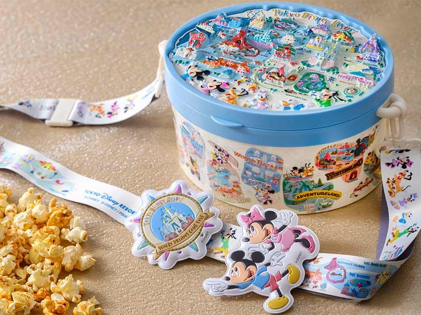 A themed Tokyo Disney Resort map popcorn bucket with colorful designs and characters, accompanied by a lanyard and character cutouts. Some popcorn is scattered beside the container.