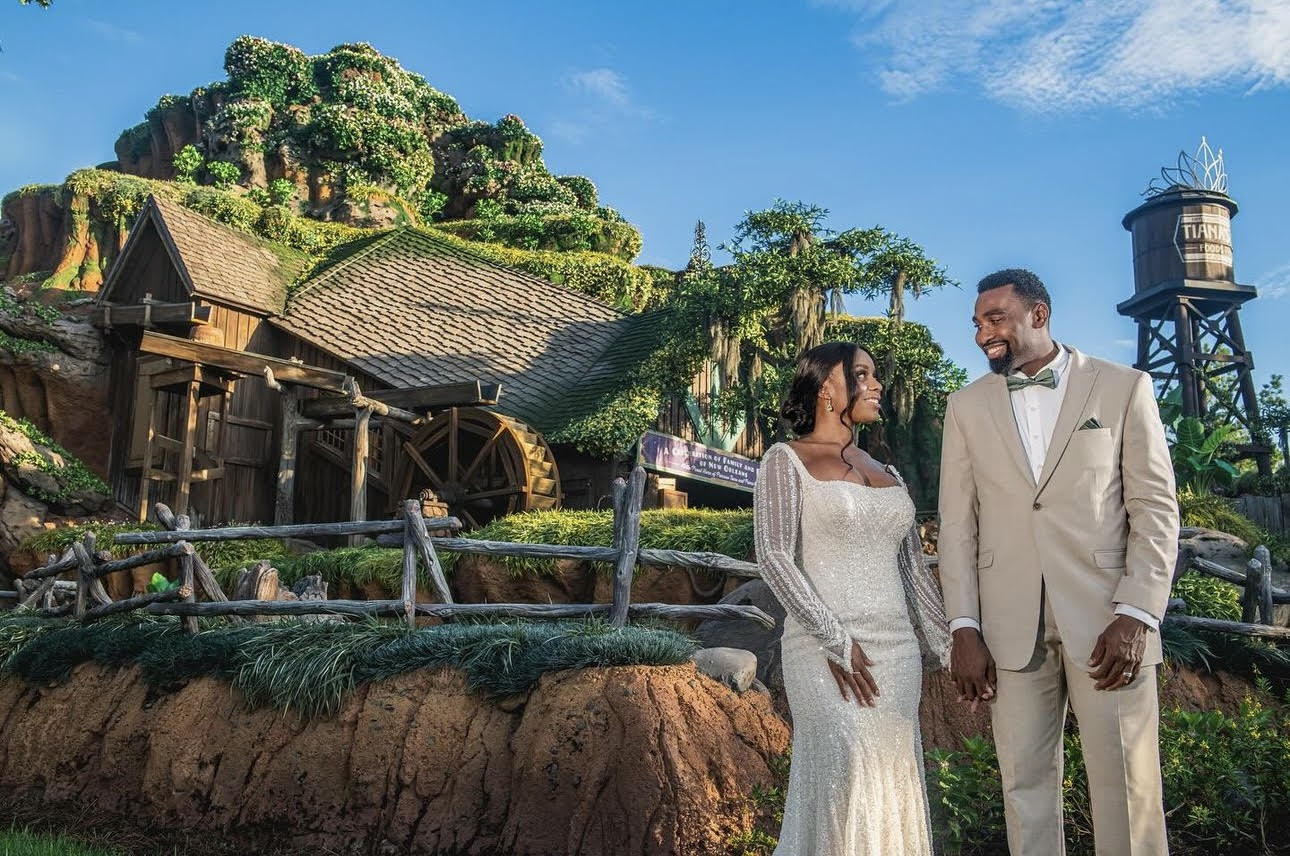 A couple in formal attire, with the woman in a white gown and the man in a beige suit, stand smiling in front of a rustic, green-covered building under a bright blue sky - an enchanting moment perfect for Disney's Fairy Tale Weddings.
