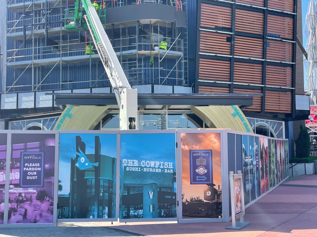 Construction site with a crane and building facade. Temporary walls display colorful placards, including signs that read, "PLEASE PARDON OUR DUST" and "THE COWFISH SUSHI BURGER BAR.