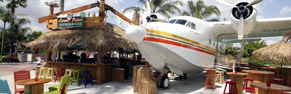Outdoor bar with colorful chairs and straw huts, featuring a large seaplane on display under a partly cloudy sky. Palm trees in the background enhance the tropical theme, evoking the laid-back vibes of a Jimmy Buffett paradise.