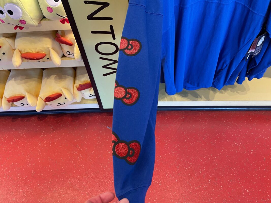 A hand is holding the sleeve of a blue Hello Kitty Spirit Jersey adorned with three red and black printed designs, while plush toys are displayed on shelves in the background.