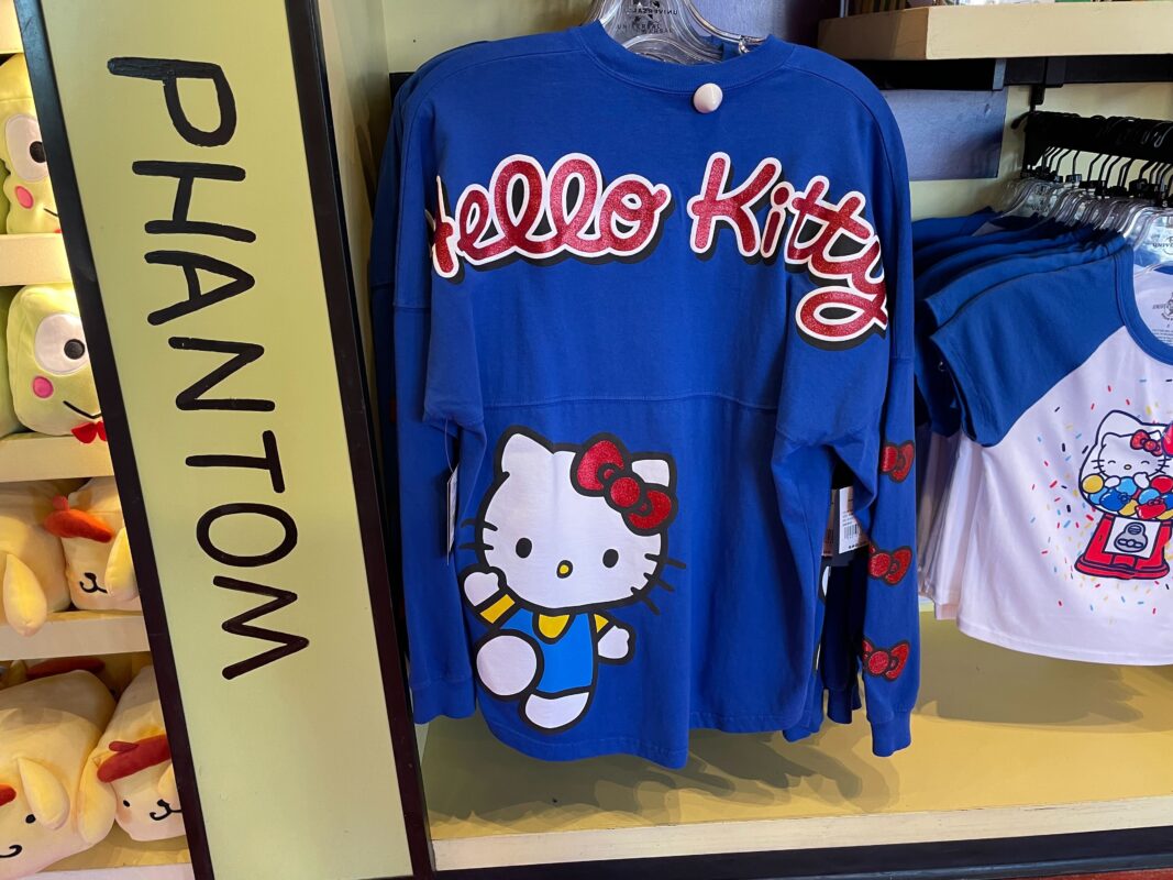 A blue long-sleeve "Hello Kitty" Spirit Jersey with red and white lettering and a graphic of Hello Kitty hangs on a rack near other merchandise in the Universal Orlando Resort store.