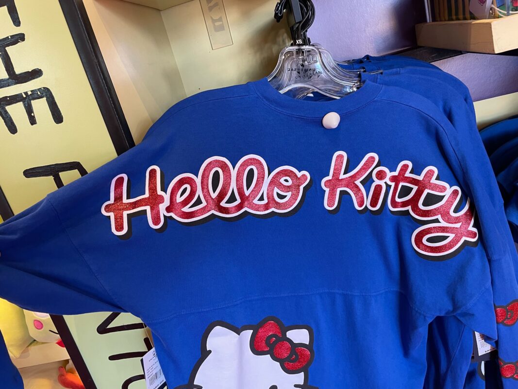 A blue Hello Kitty Spirit Jersey with "Hello Kitty" written in red and white letters on display at Universal Orlando Resort. The shirt features a partial image of the Hello Kitty character's face with a red bow.