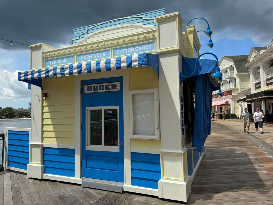 A small boardwalk food kiosk with a blue and white awning offering lemonade, corn dogs, and cotton candy. The sign above the window reads "ORDER." The sky is partly cloudy.