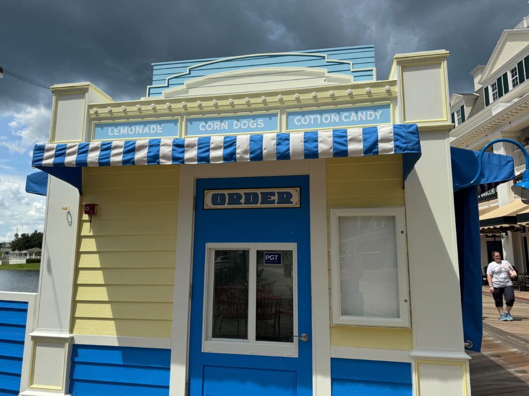 A small yellow and blue food stand with signs for lemonade, corn dogs, and cotton candy. The stand features a striped awning and an “Order” window in the center.
