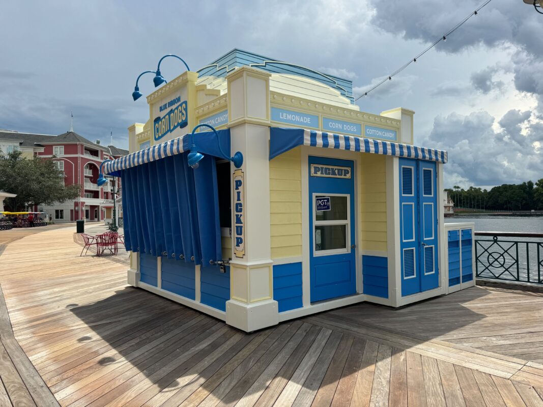 A small, blue and yellow food stand on a boardwalk offers lemonade, corn dogs, and cotton candy. The stand has a "Pick Up" window and is flanked by striped blue awnings.