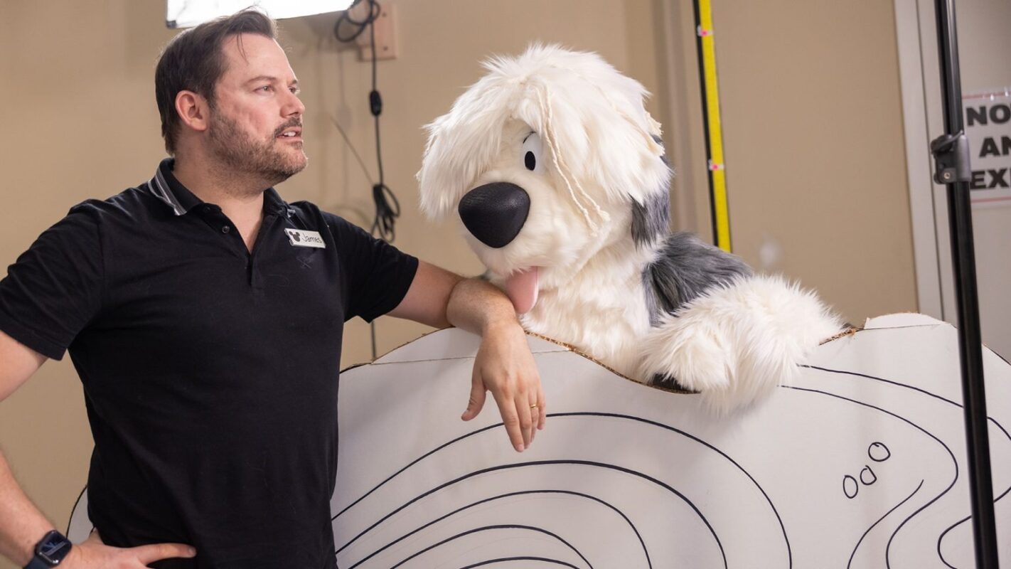 A man stands next to an oversized fluffy dog mascot prop, leaning on a large piece of white card featuring contour lines. The backdrop is an indoor space with a beige wall and some equipment visible.