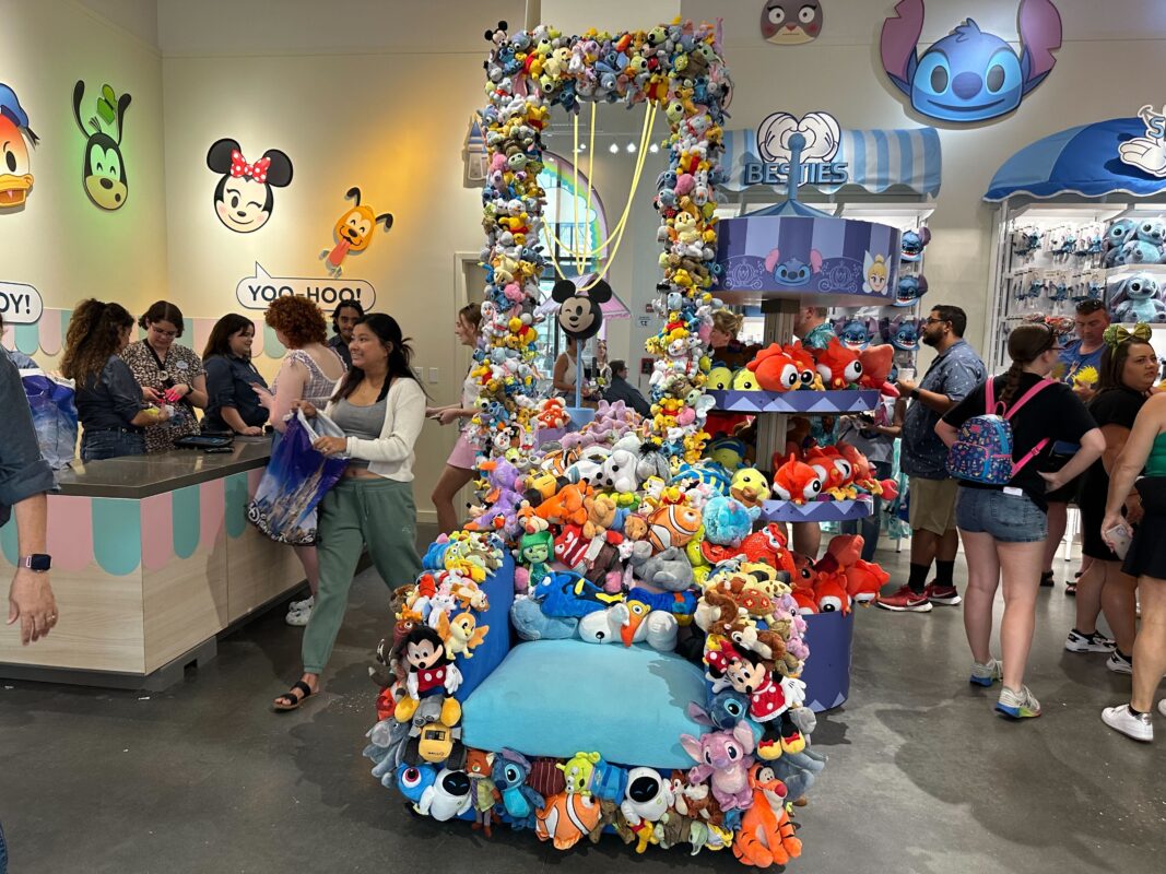 A store displays a variety of stuffed toys around a throne-like chair adorned with plushies. People shop and browse other merchandise nearby. Colorful illustrations decorate the walls.
